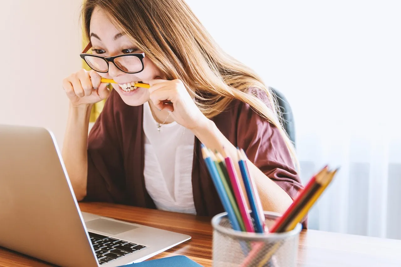 Woman in glasses holding a pencil while reviewing her laptop screen in a bright creative workspace.