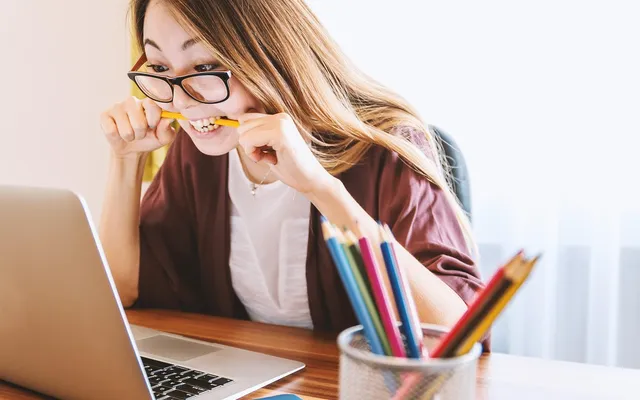 Woman in glasses holding a pencil while reviewing her laptop screen in a bright creative workspace.